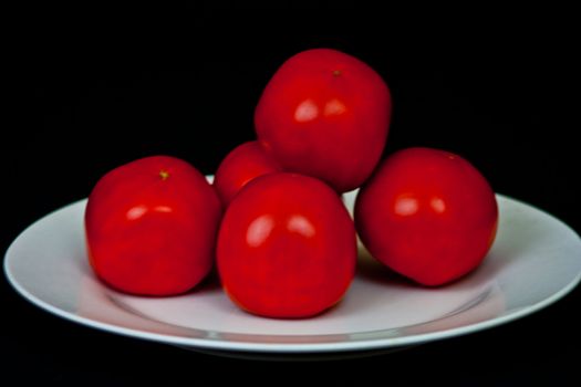 A pile of red tomatoes on a white plate on a black bacground.