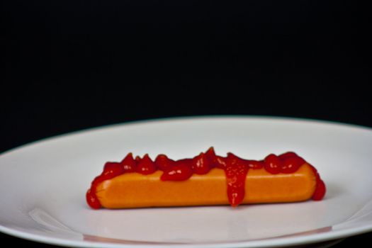 Hot dog with Ketchup on top sitting on a white plate on a black background.