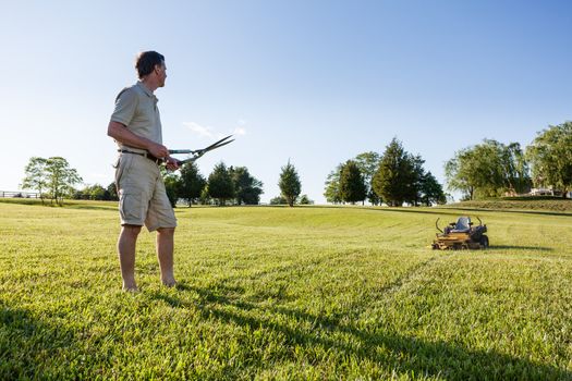 Challenging task of cutting large lawn with grass shears by hand with mower in background