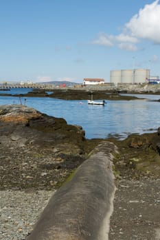 A concrete pipe leads across a beach to an area of water with a boat and an industrial complex in the background.