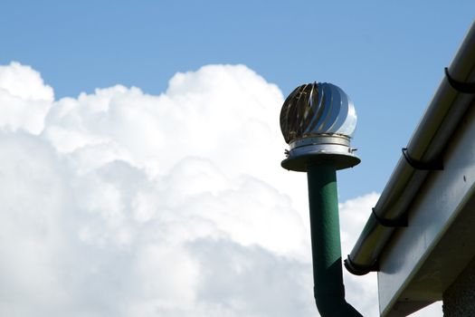 A wind powered rotor vent system with duct by a roof gutter with a blue sky and bulbous white cloud in the distance.