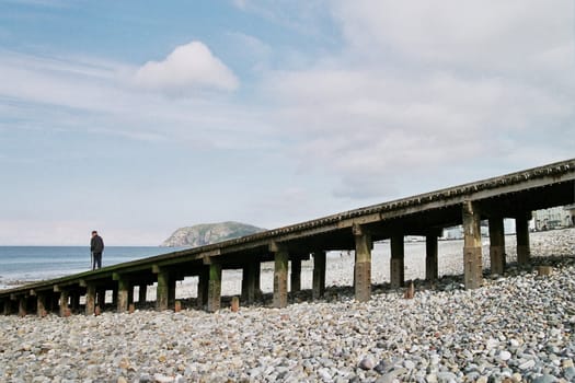 A senior man stands on the end of a wooden slipway looking out to sea.