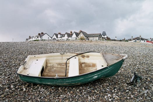 An old wooden rowing boat painted green rests on a pebble beach with buildings in the distance.