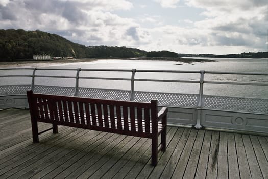 A wooden brown stained bench on boards with a railing and the sea, trees and sky in the distance.