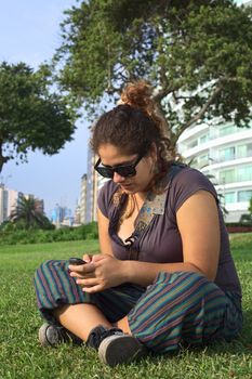 Beautiful young Peruvian woman text messaging on mobile phone in park (Selective Focus, Focus on the face and the hands of the woman)