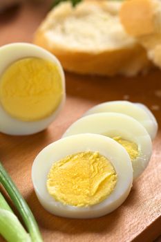 Fresh hard boiled egg cut in slices with scallion beside and baguette in the back on wooden cutting board (Selective Focus, Focus on the upper edge of the egg yolk of the first slice)