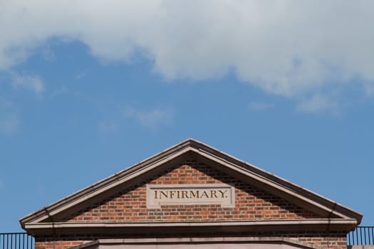 A triangular shaped structure, brick wall and roof coping stones with a sign 'INFIRMARY' written on it against a blue sky with cloud.