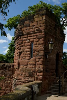 An historic wall tower built from sandstone with steps, doorway and trees against a blue sky.