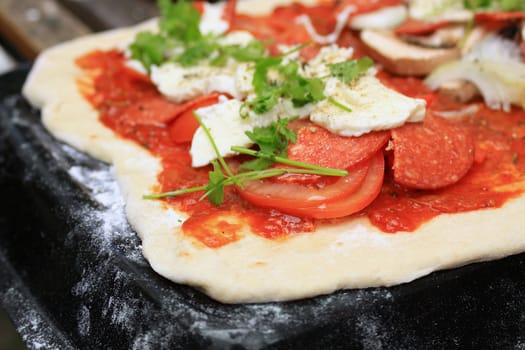 A homemade pepperoni sausage, mozzarella cheese, tomato, mushroom and onion pizza, with fresh coriander, set on a homemade pizza dough base with a homemade tomato sauce. Set on a flowered baking tray ready for the oven. Set on a landscape format with copy space available.