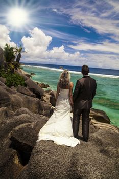 Bride and groom standing holding hands on rocks looking out over the ocean at the seashore under tropical sunshine