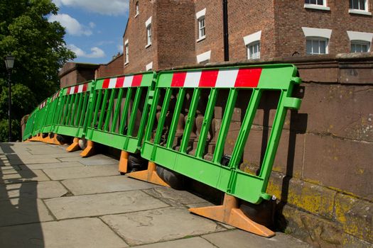 An open walkway with green plastic barriers, with red and white warning symbols leaning against a stone wall.