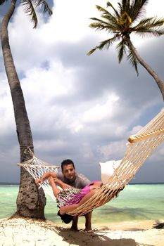 Woman relaxing in a hammock strung between palm trees on a tropical beach chatting to a handsome young man