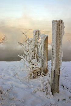 winter misty landscape on the river with the snow-bound blasted fence