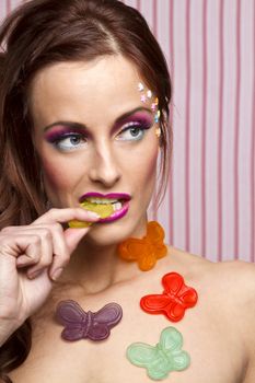 Young woman with colorful makeup and star candy glued to her face and gummy butterfly glued to her body, sucking on the wing of a yellow butterfly