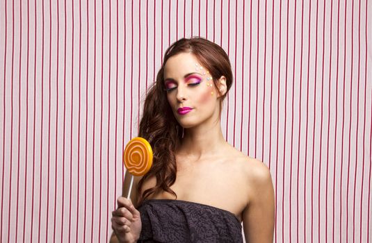 Young woman with colorful makeup and star candy glued to her face, with her eyes close holding an orange lollipop