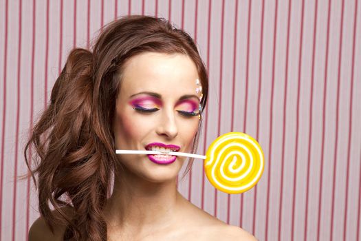 Young woman with colorful makeup and star candy glued to her face, holding an orange lollipop by the stick between her teeth