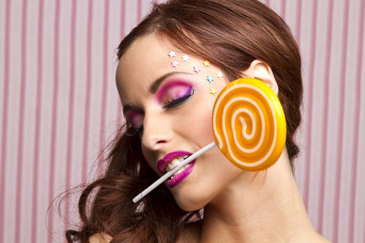 Young woman with colorful makeup and star candy glued to her face, holding an orange lollipop by the stick between her teeth