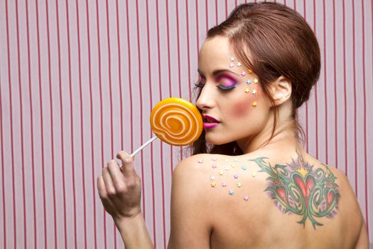 Young woman with colorful makeup and star candy glued to her face, licking an orange lollipop