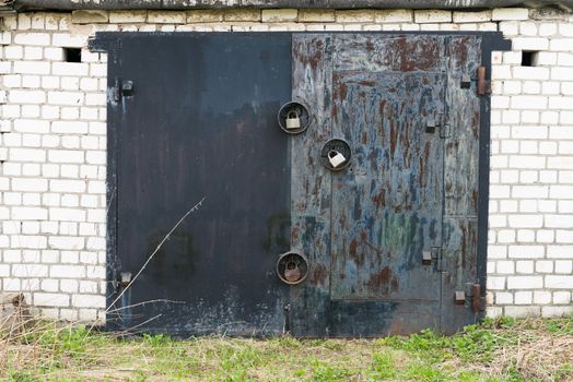 Aged closed black metal garage gate with three big locks on white brick wall