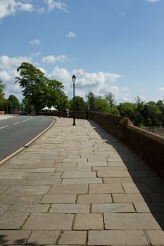 A random flagstone pavement leads into the distance next to a wall with a lamppost, people and a road and trees against a blue sky.