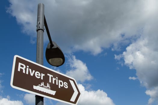 A brown sign with  a white border and the symbol for a boat and the words 'RIVER TRIPS' attached to a lamppost against a blue sky with cloud.