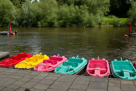 A row of multicoloured boats lined up next to a river bank pavement with trees on the far shore.