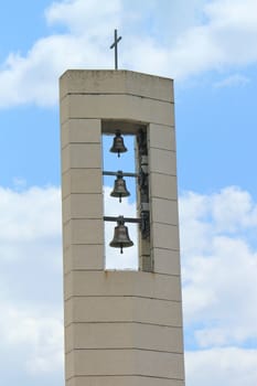 Three bells in a modern bell tower with a cross upon and cloudy sky