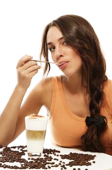 young woman with spoon in her mouth sitting at a table wit latte macchiato coffee on white background