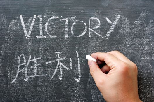 Victory - word written on a blackboard with a Chinese translation, with a hand holding chalk