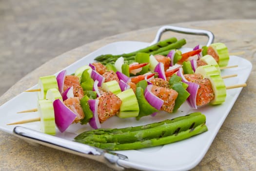 Salmon cubes and Shrimp complimented with vegetables on skewers. Red onions, zucchini, green peppers and asparagus. On white modern plate placed on concerete countertop.