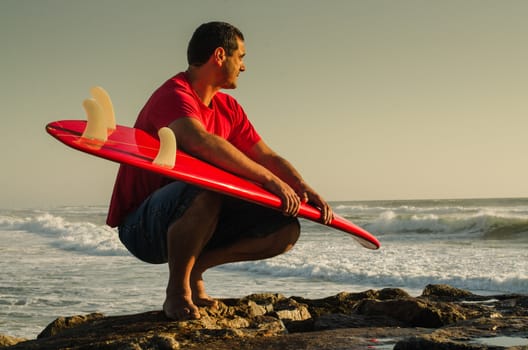 A surfer watching the waves sitting down with his arms around his surfboard.