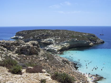 This is the magnificent island of rabbits, in Lampedusa. The water is crystal clear and the sand is white. The rocks are silhouetted against the blue sea and the sky is clear. This area is protected reserves.