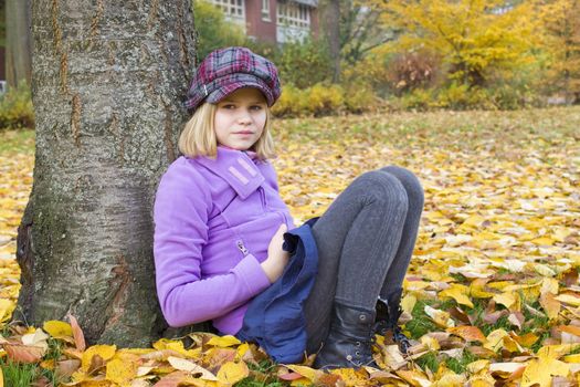Full length portrait of a little girl sitting under tree in autumn park 