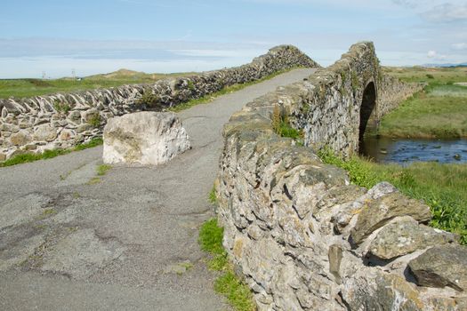 An old bridge with a boulder blocking vehicle accesss with walls and a path leading over a river into the distance with a blue cloud sky beyond.