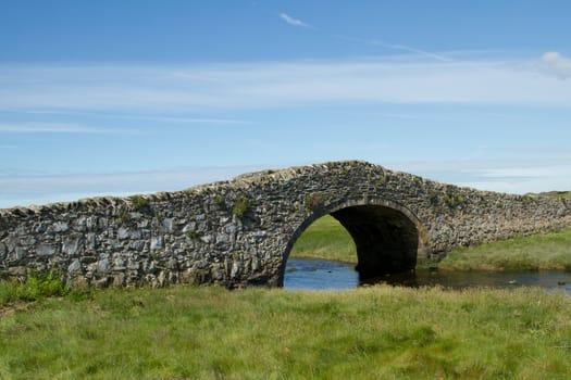 An historic narrow stone built bridge crossing a river with green grass and a blue cloudy sky in the distance.