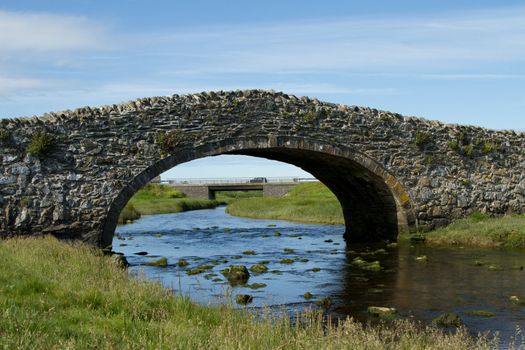 An historic narrow stone bridge crosses a river in the foreground with a view of a new bridge under the arch with a car travelling across against a blue cloudy sky.