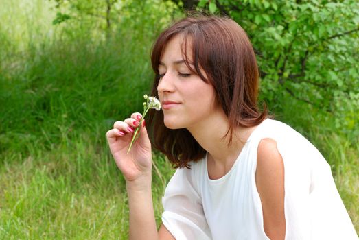 portrait of young caucasian brunette woman in white dress smelling a flower over natural green background