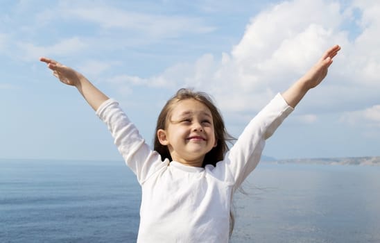 little cute girl standing arms outstretched against a background of blue sea and sky with clouds