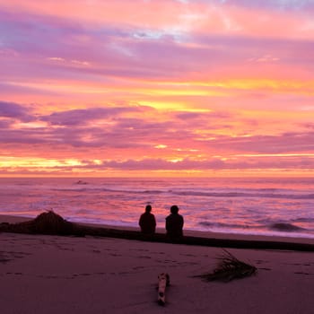 Romantic couple sitting close together on the sand enjoy a spectacular beach sunset over the ocean