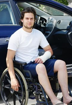 Young man with plaster cast after an accident in wheelchair arriving or leaving the hospital, car with opened door in the background.