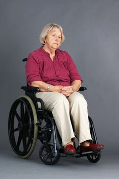 Sad or depressed senior woman in a wheelchair, looking down, studio shot over grey background.