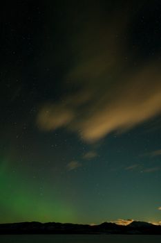 Northern lights (aurora borealis) and drifting clouds lit by rising moon above and behind snowy mountain range in winter.