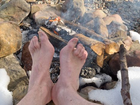 Bare feet of one person are warming at a Campfire outdoors at wintertime with patches of snow.