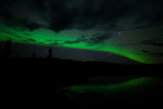 Night Sky Stars, clouds and Northern Lights mirrored on calm lake in Yukon, Territory, Canada.