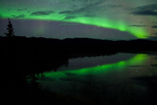 Night Sky Stars, clouds and Northern Lights mirrored on calm lake in Yukon, Territory, Canada.