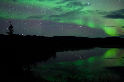 Night Sky Stars, clouds and Northern Lights mirrored on calm lake in Yukon, Territory, Canada.