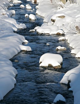 A sunny day view of a small mountain river with fresh snow on the banks.