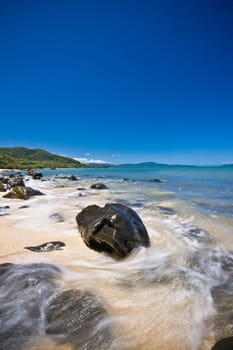 Water flowing over rocks on a beautiful sandy sunlit tropical beach with focus to a single rock standing clear of the surf