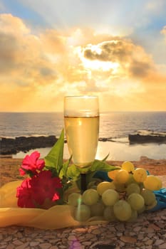glass of white wine, grapes and flowers on a background of sea and sky at sunset