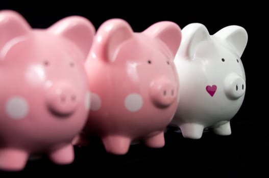 Three little piggy banks lined up. Unique white heart piggy in focus against black background